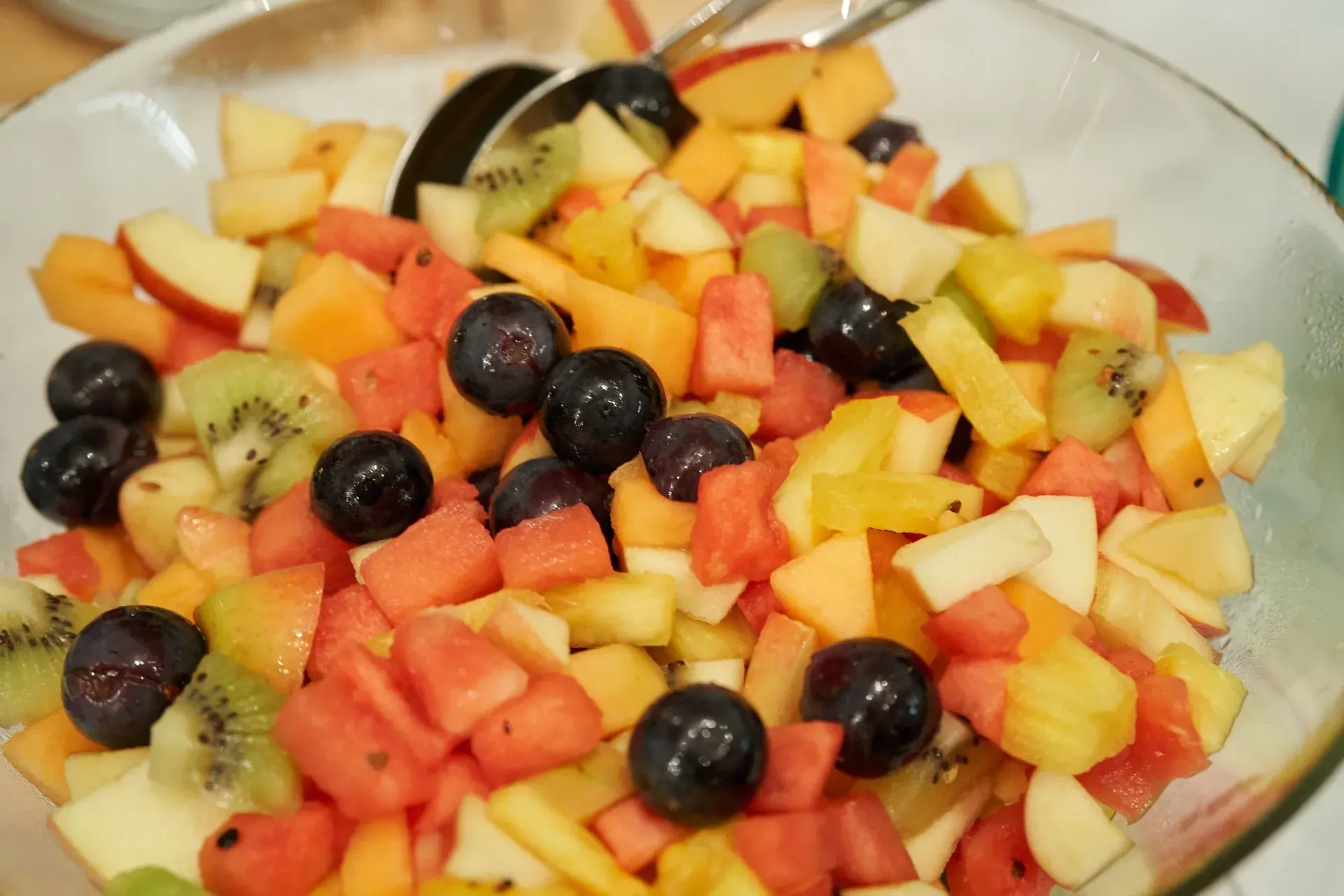 A glass bowl of fresh-cut tropical fruit salad with serving spoons