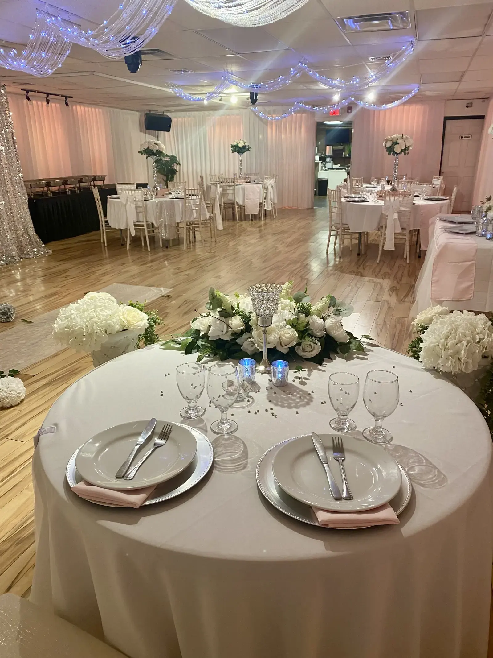 A reception set with round tables in blush and white, chiavari chairs, a sweetheart table, and chafing dishes on the buffet