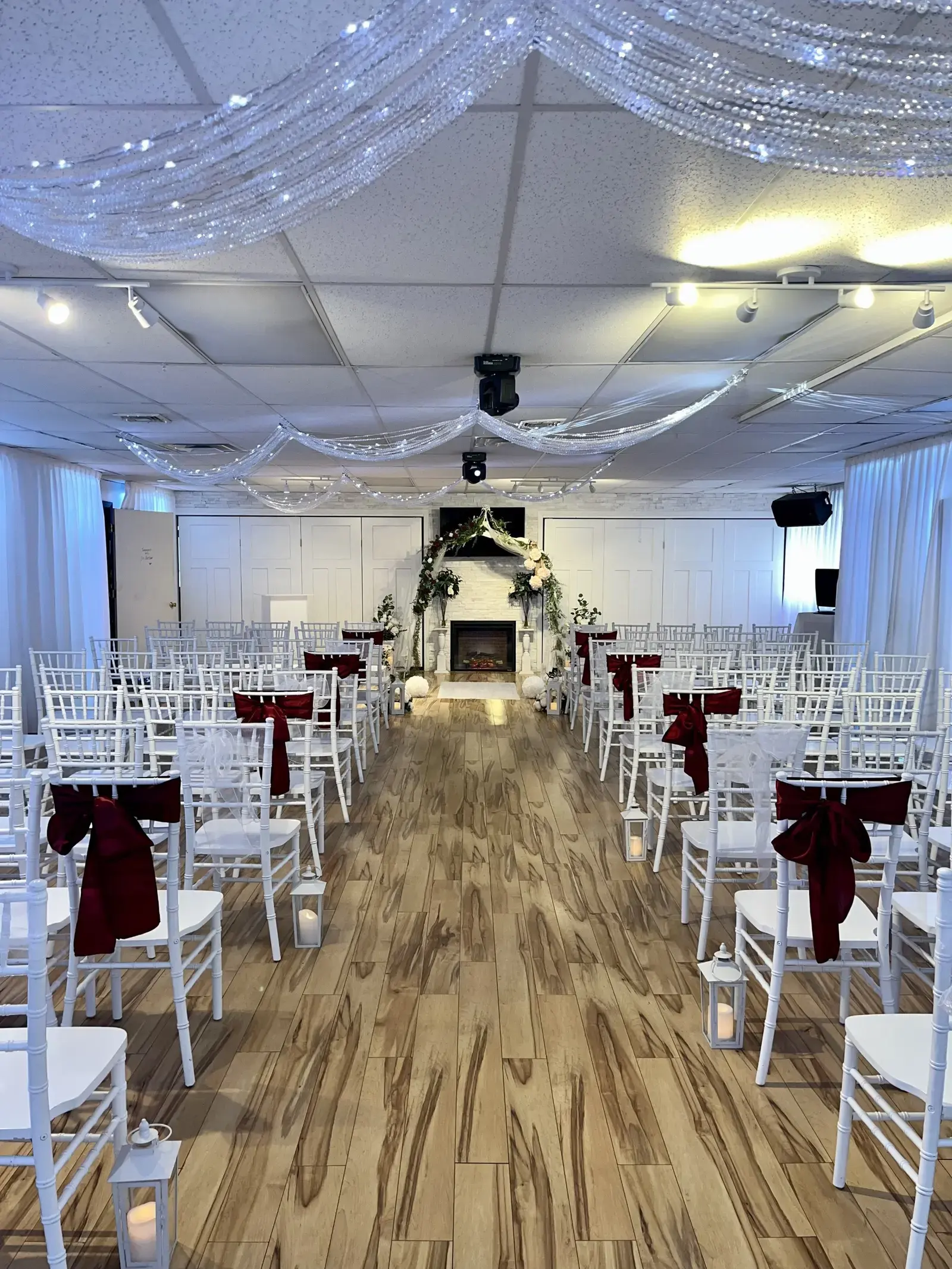 The room set for a ceremony — white chiavari chairs with burgundy sashes lining an aisle toward a floral arch at the fireplace